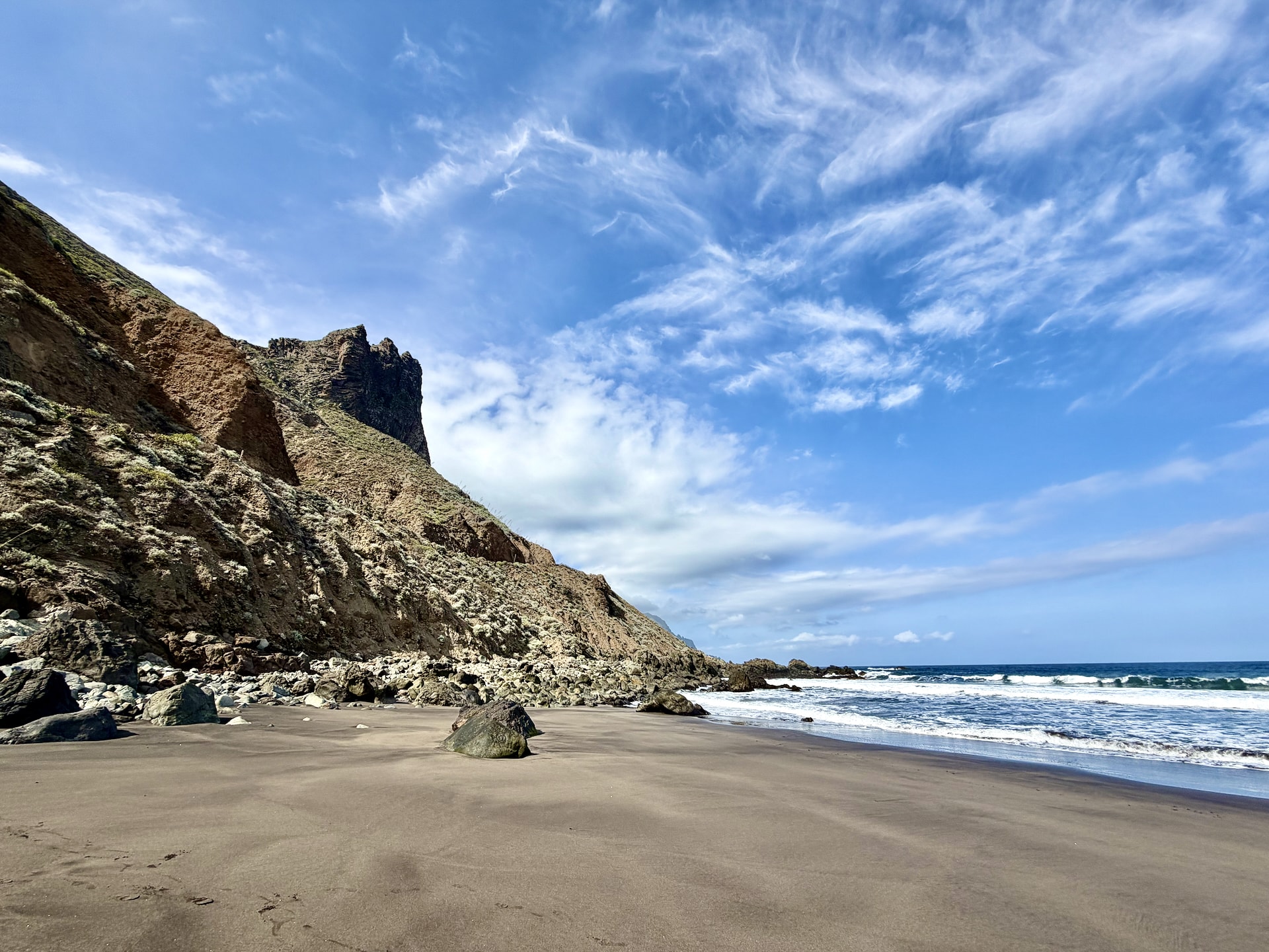 Dark sand beach below steep rocky cliffs at Playa del Roque de las Bodegas in the Anaga mountains, with waves rolling in under a blue sky