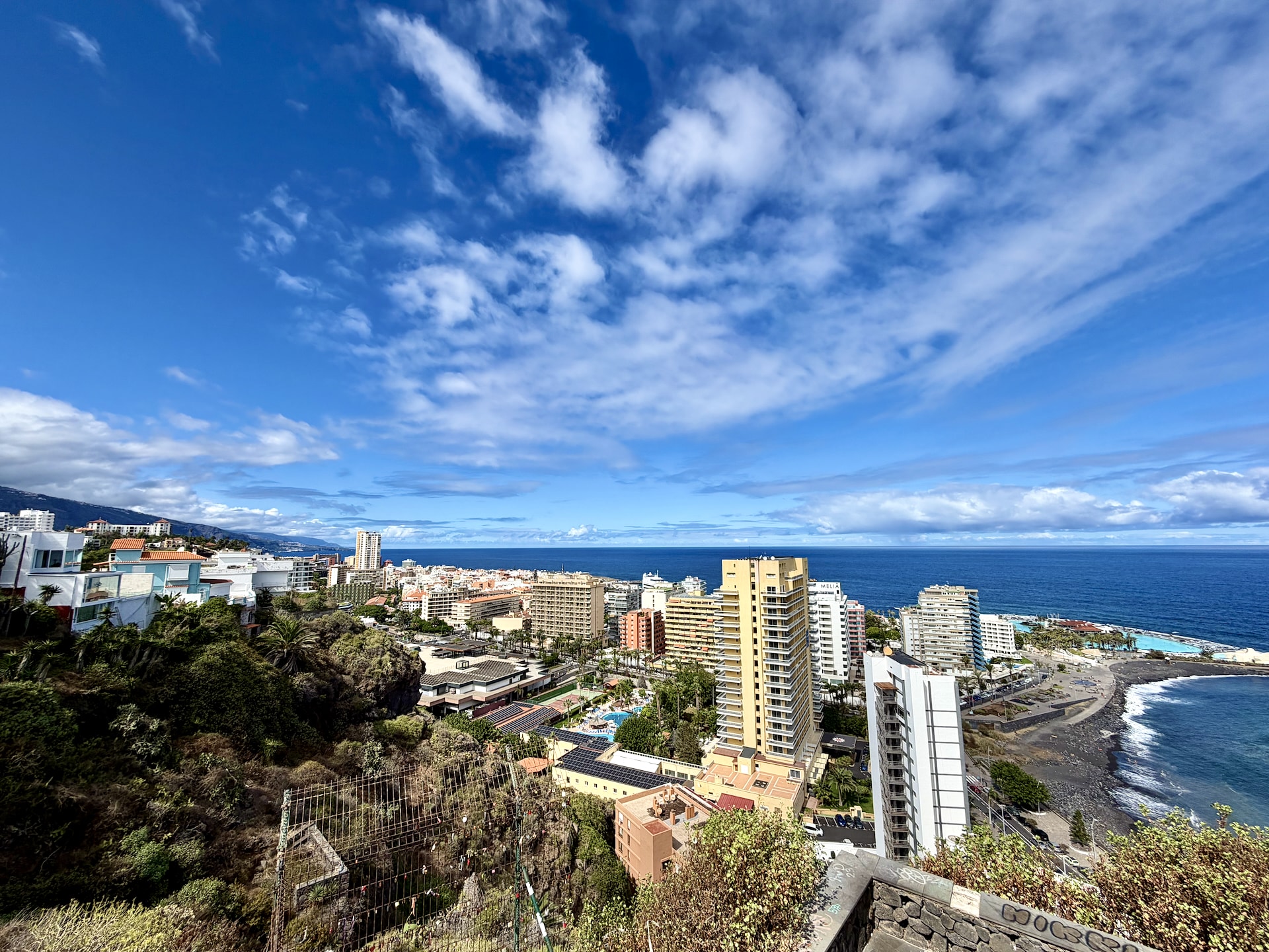 Hotels and apartment towers of Puerto de la Cruz seen from Mirador de La Paz, with the Lago Martiánez pools and a black sand beach on the right
