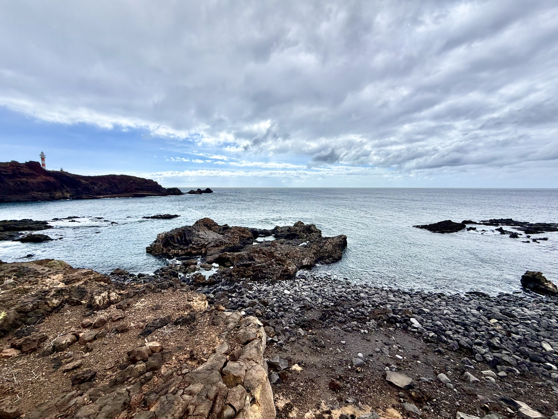 Black volcanic shoreline at Punta de Teno with the lighthouse far off on the headland under a cloudy sky