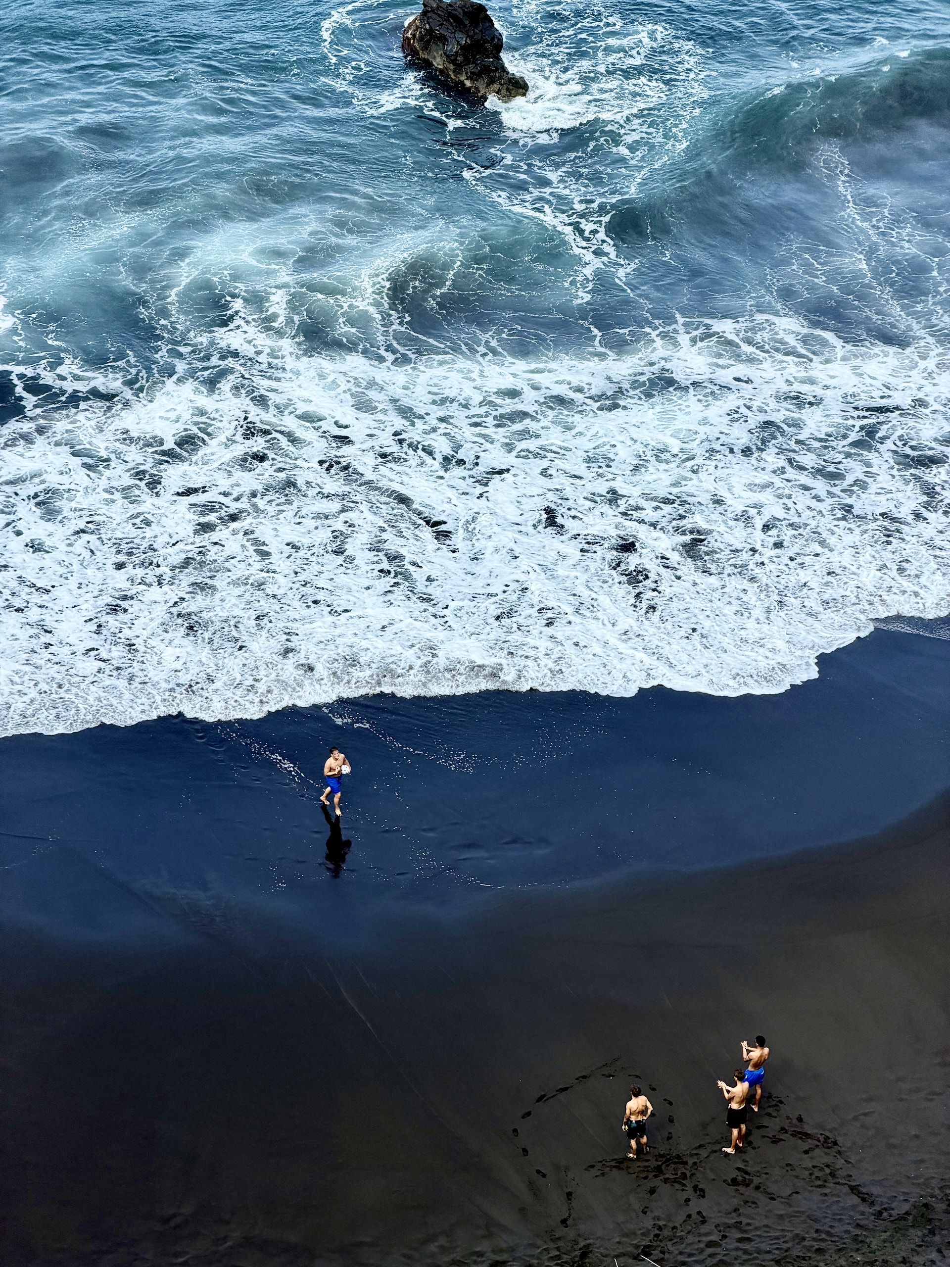 Rescuing a ball from the waves at El Bollullo Aerial view of El Bollullo: a man carrying a ball back from the surf while three others wait for him on the black sand