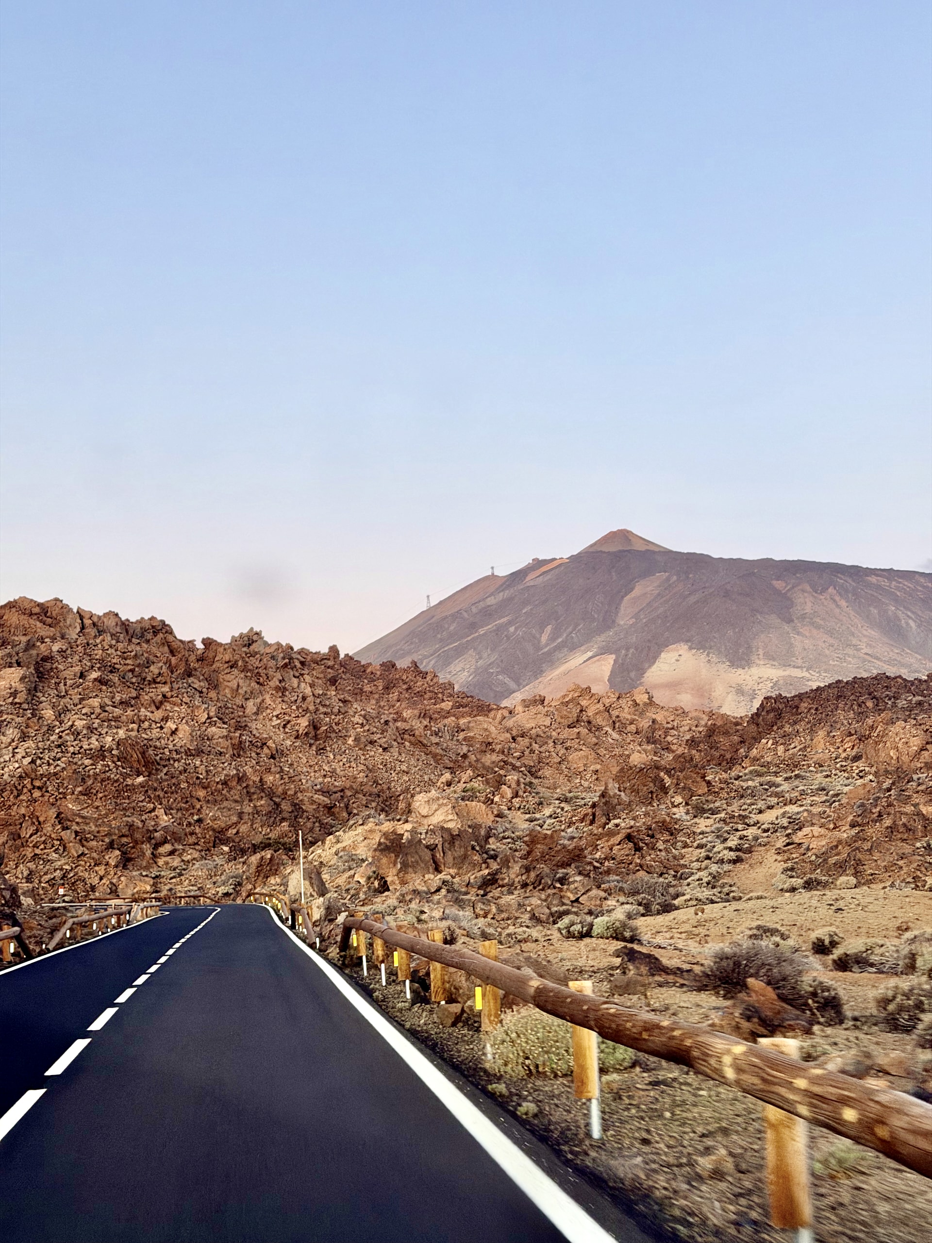 An empty road with a wooden guardrail winding through red-brown lava fields towards Mount Teide in the early morning