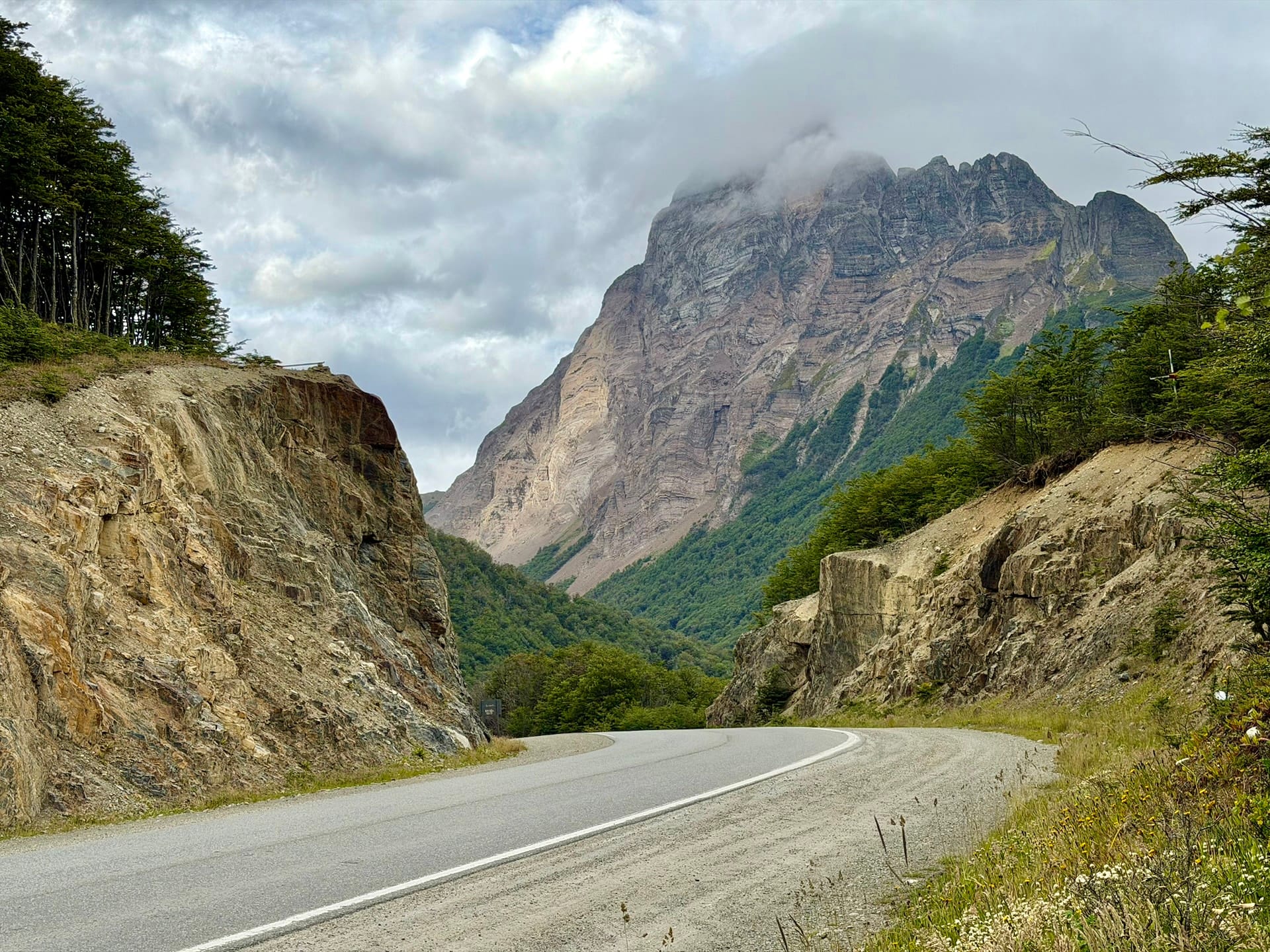 Ruta 3 curving through rock cuttings east of Ushuaia beneath a steep cloud-capped mountain