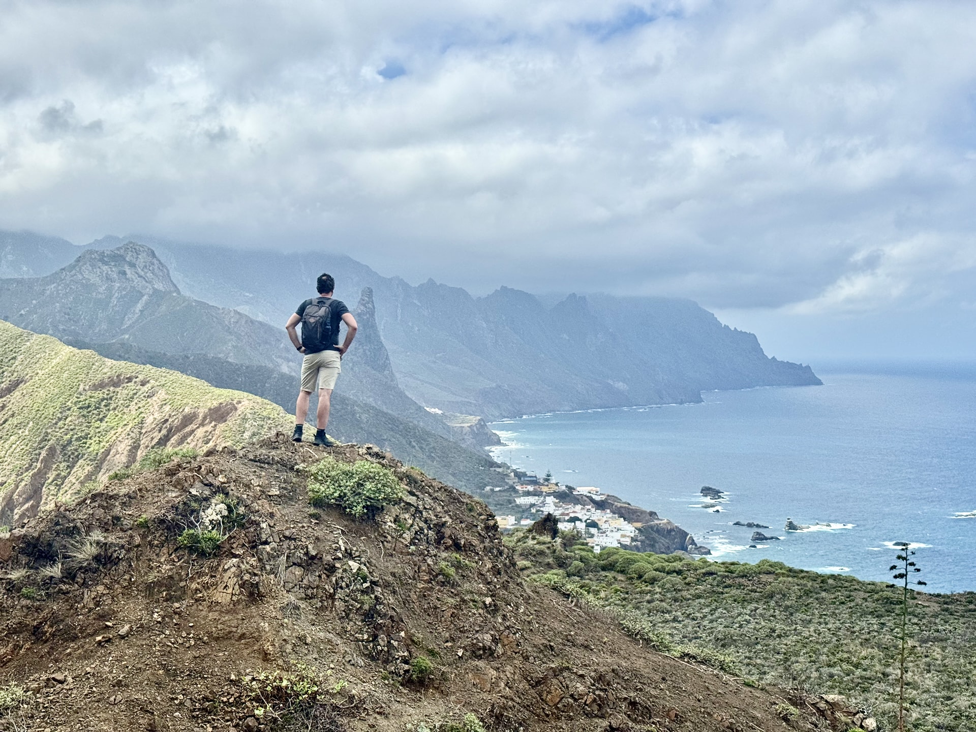 Laszlo standing on a ridge above the Anaga coast, looking out over steep green mountains, a coastal village and the Atlantic