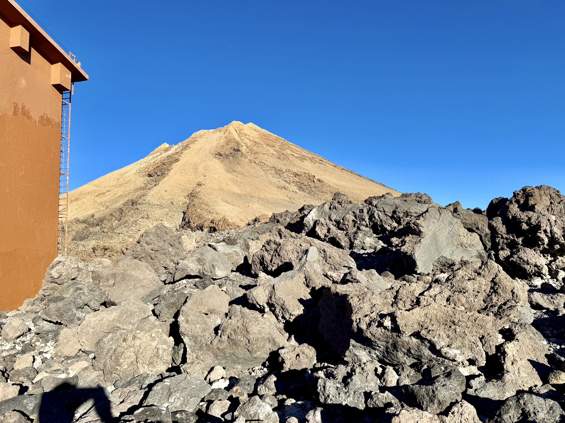 The pale summit cone of Mount Teide above dark lava rocks, seen from beside the upper cable car station at La Rambleta