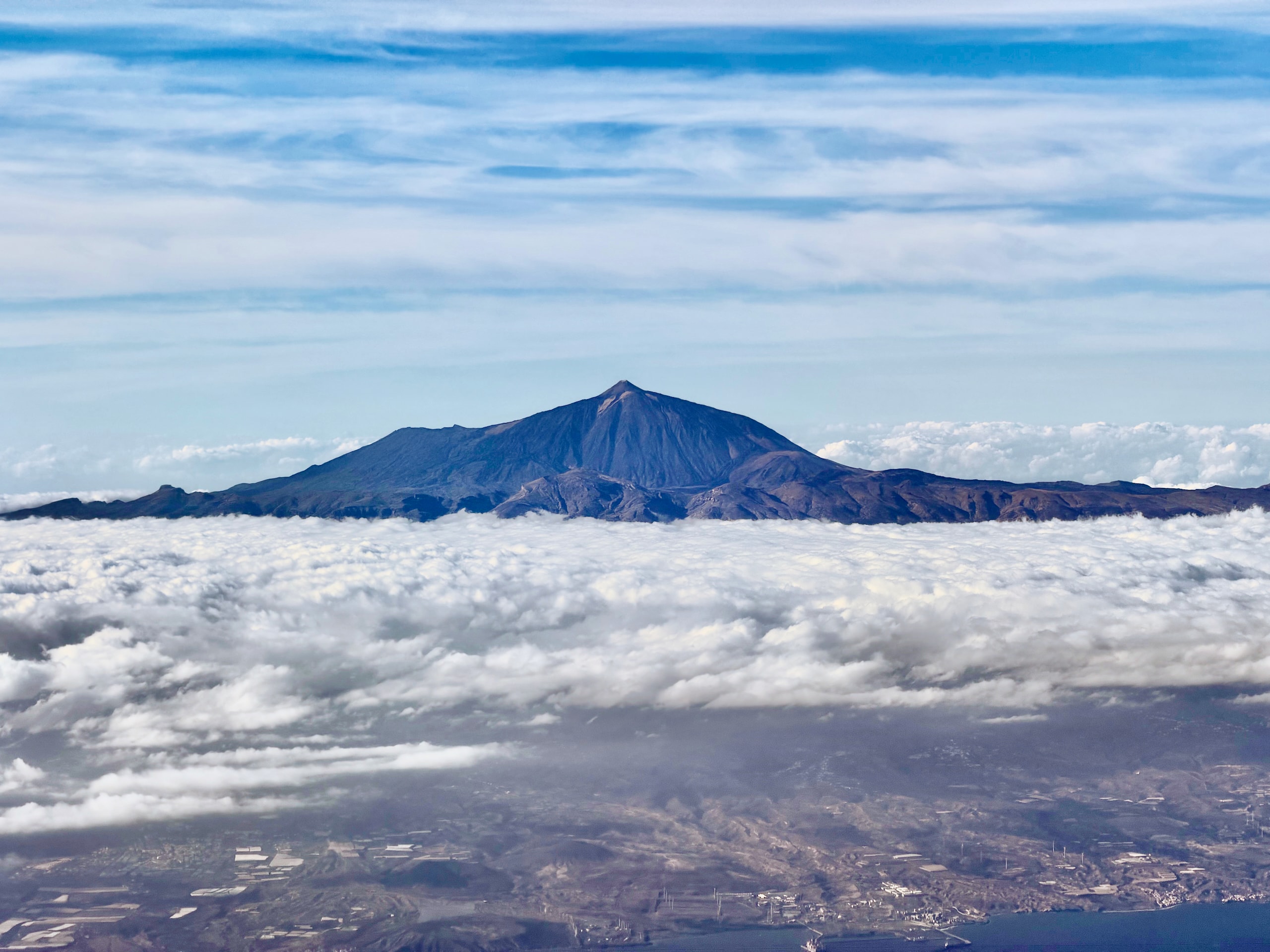 Mount Teide rising above a sea of clouds over Tenerife, seen from a plane window, with the island's coast visible below the cloud layer