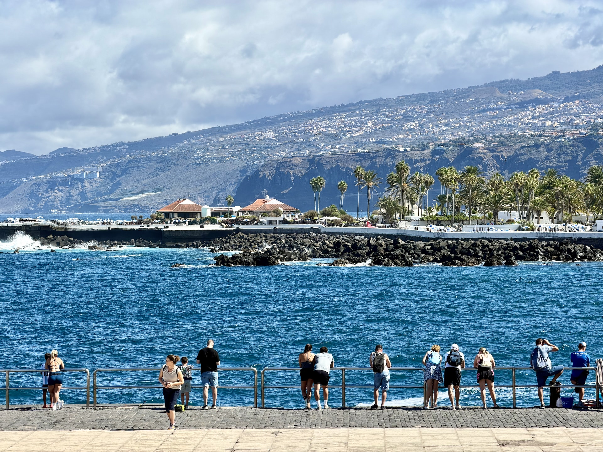 Tourists lining a railing by the old harbor in Puerto de la Cruz, looking out at the sea, with the green hills of the north coast behind