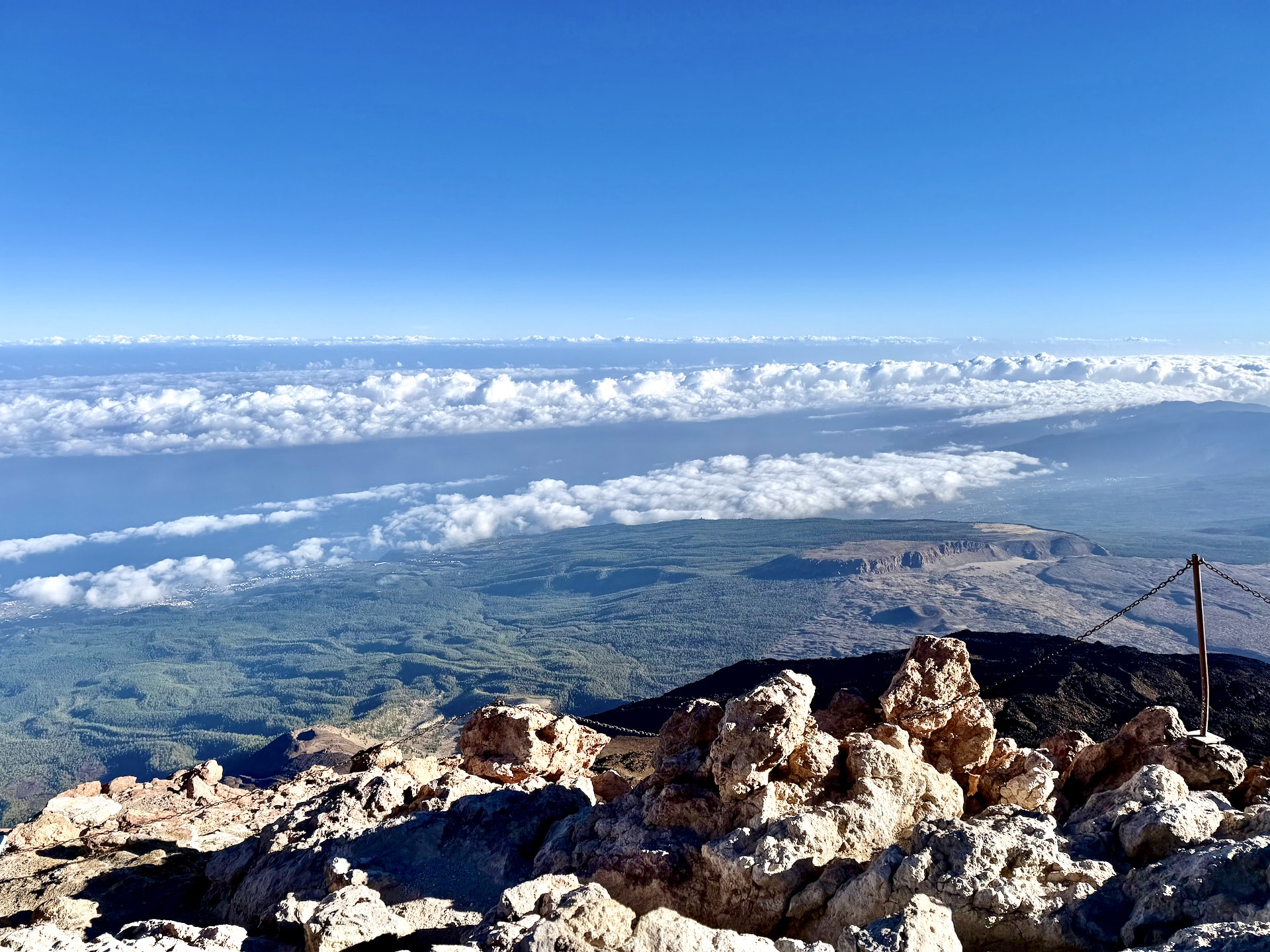 View north from the summit of Mount Teide over light-colored rocks to a band of clouds below, the green northern slopes of Tenerife and the Atlantic Ocean