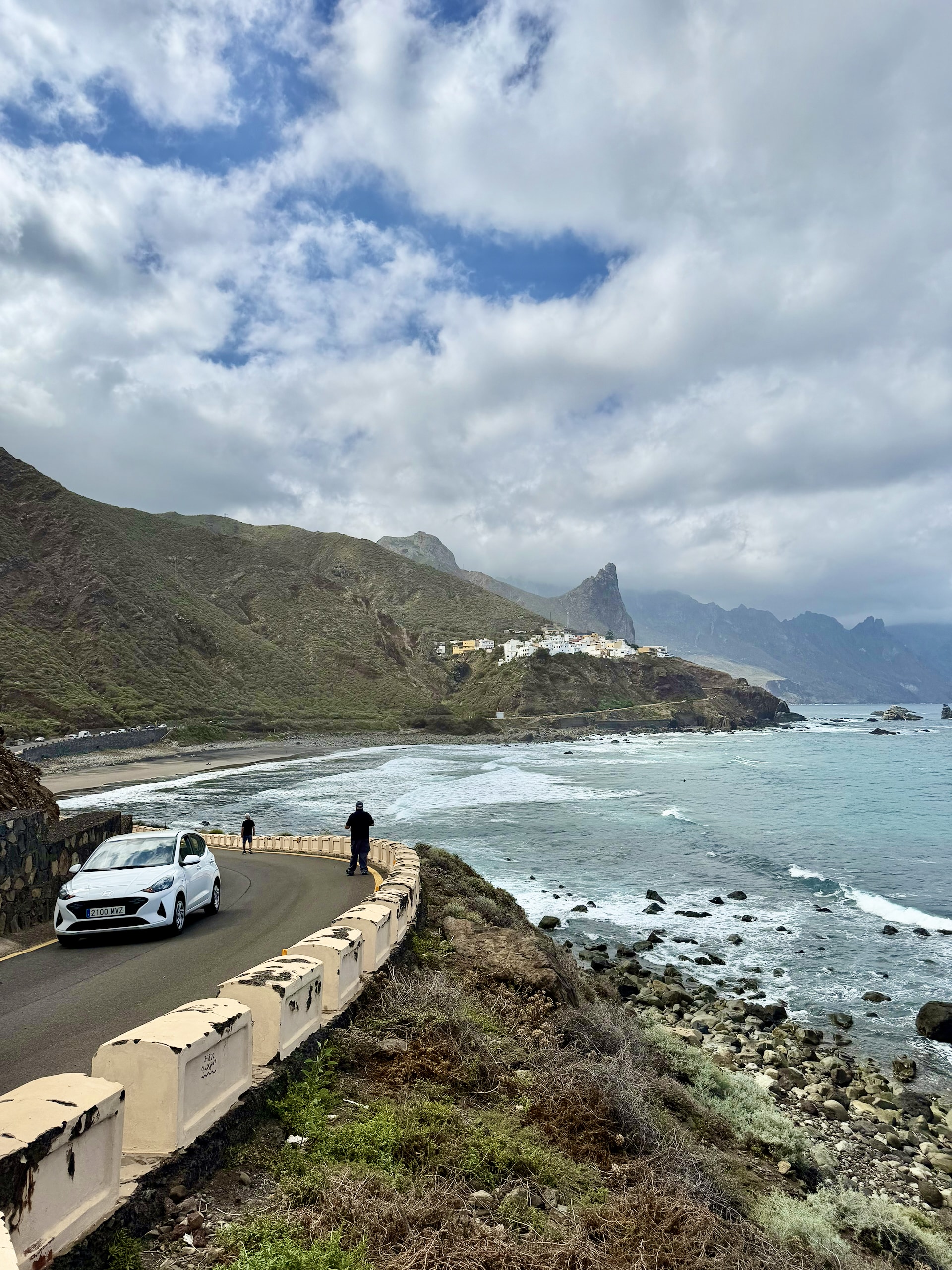 Parked car and people taking photos on the coastal road near Benijo, above a rocky shore, with a village and a rock spire in the distance