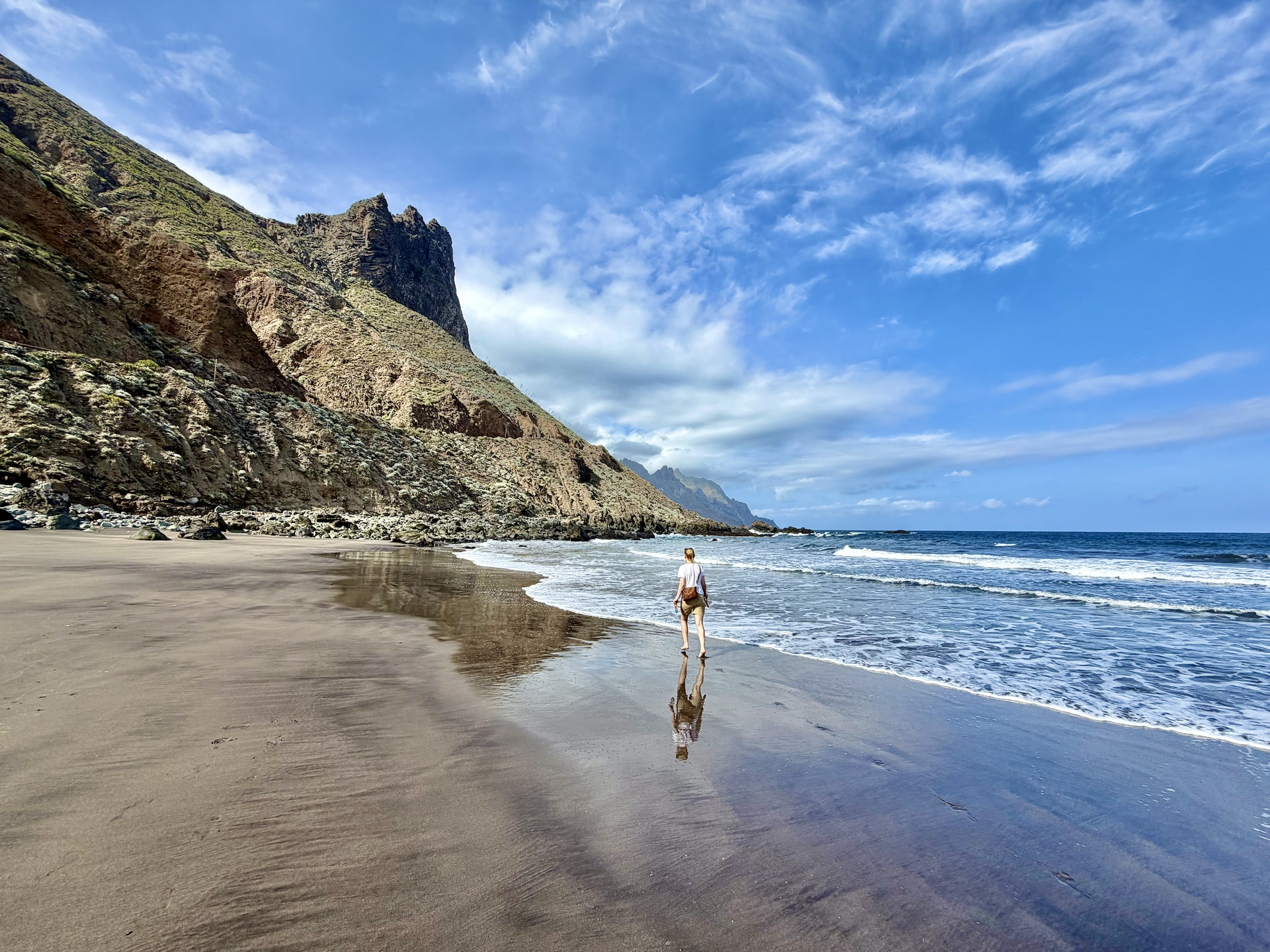 Andrea walking along the wet sand at Roque de las Bodegas beach, with steep mountains and a jagged peak behind