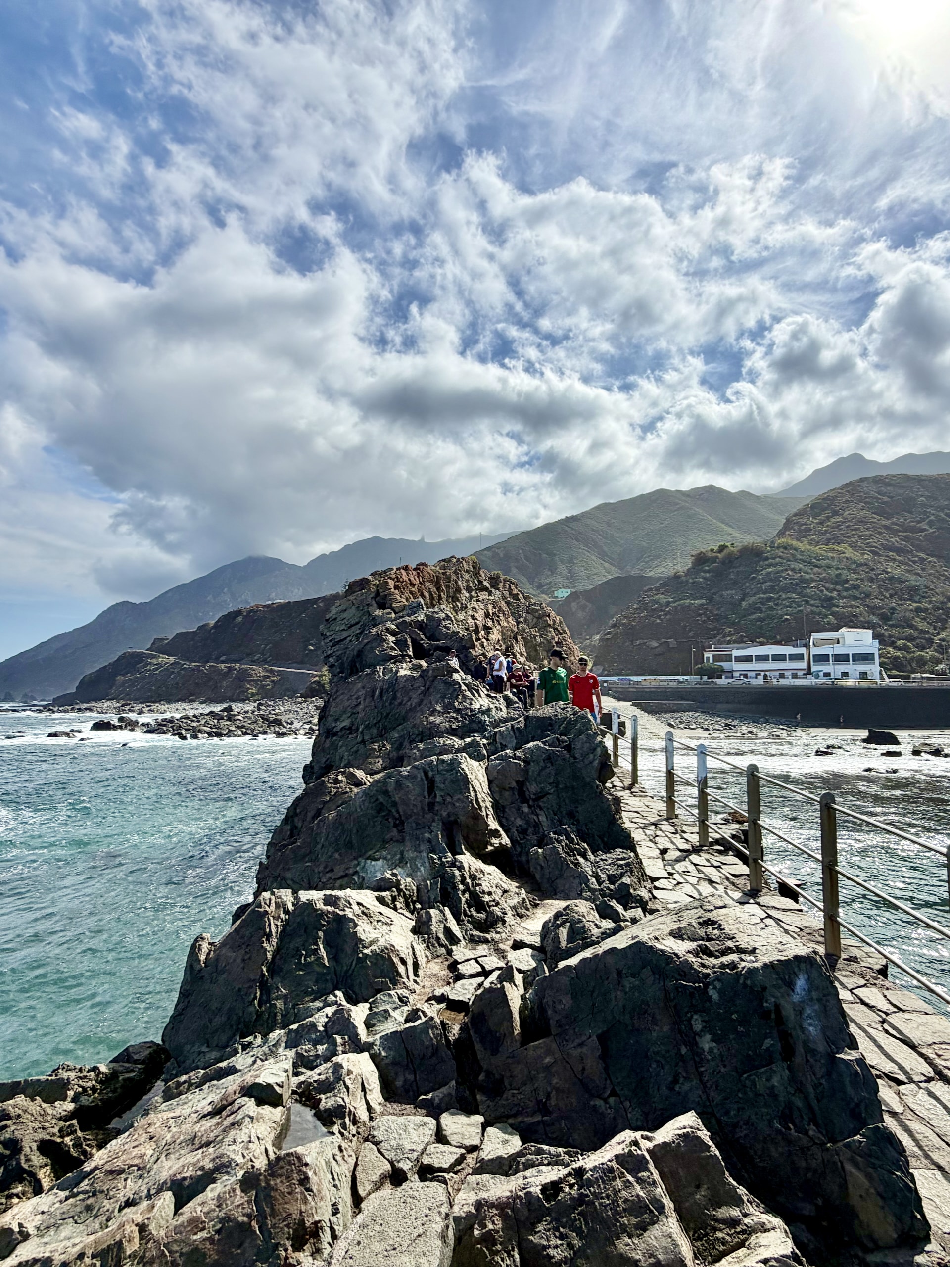 Walkway with a railing along the top of the rock at Roque de las Bodegas, with the sea on both sides and the Anaga mountains behind